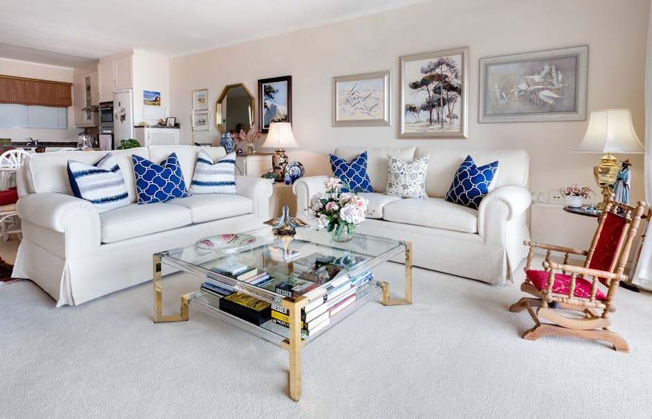 A bright, well-lit living room featuring two white upholstered sofas with blue and white patterned cushions arranged along a cream-colored wall adorned with framed artwork depicting nature scenes. In