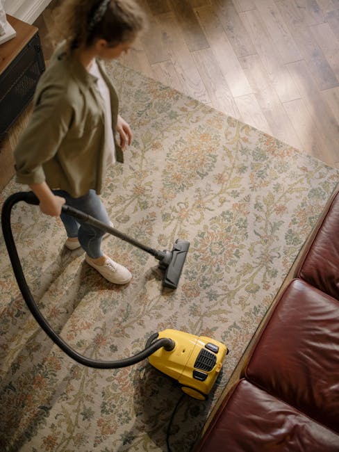 A woman using a yellow and black vacuum cleaner to deep clean a patterned area rug in a living room. The rug features intricate floral designs in shades of beige, green, and orange, laid on a wooden f
