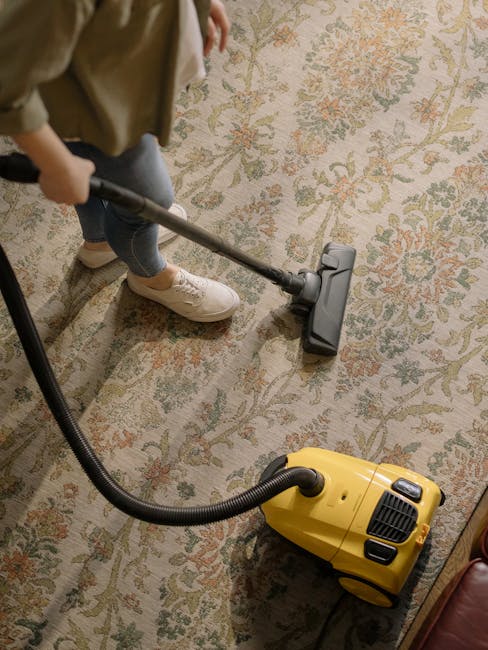 A woman using a yellow and black vacuum cleaner to deep clean a patterned area rug in a living room. The rug features intricate floral designs in shades of beige, green, and orange, laid on a wooden f